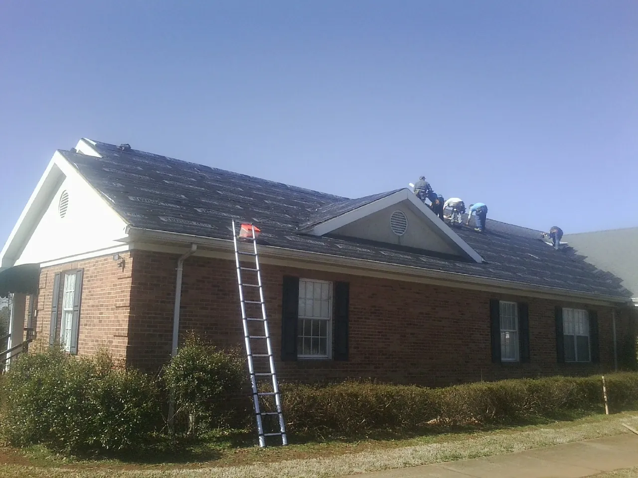 Crew tearing shingles off a brick ranch roof with a ladder set — Metal Roof Repair work by our crew in Evensville