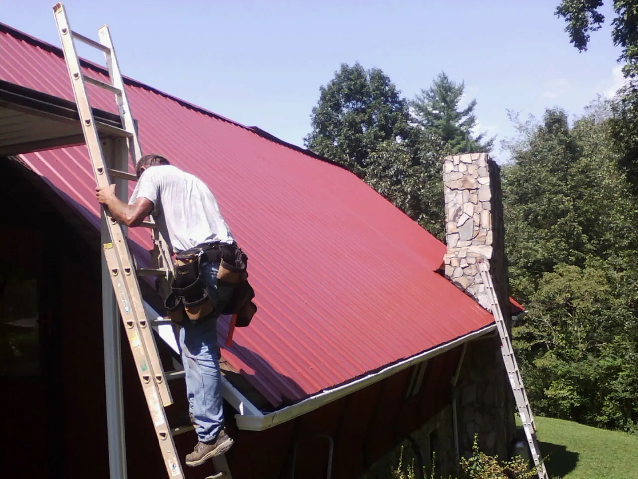 Roofer on a ladder beside a red metal roof and stone chimney — Soffit & Fascia Repair by Roofing Brothers in Evensville